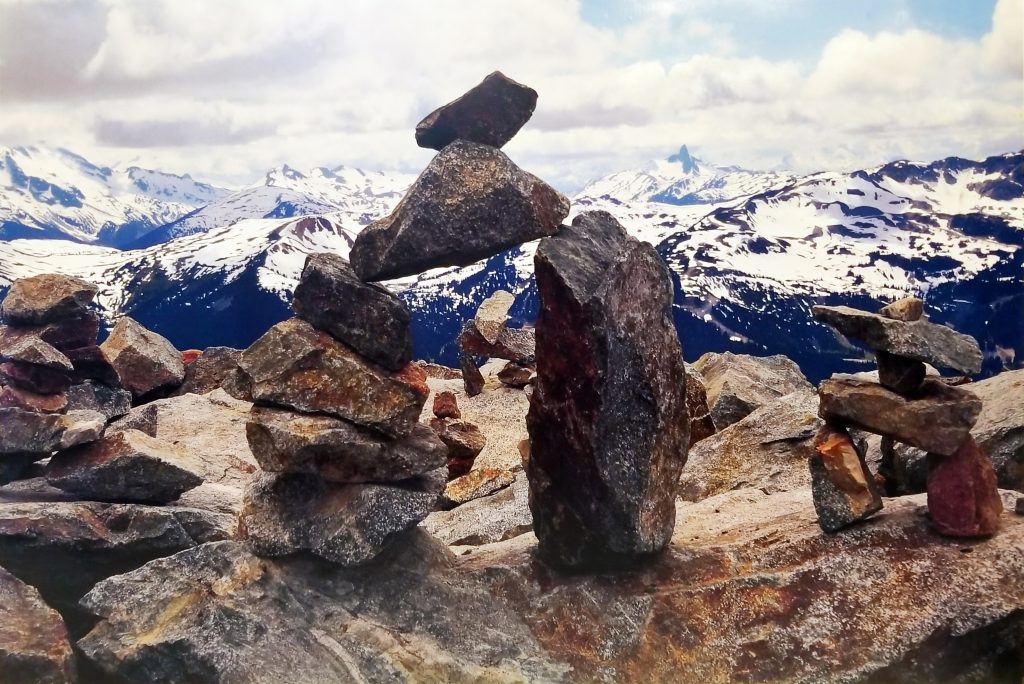 Inuksuk on rocky terrain with snow-capped mountains and a blue sky with rolling clouds overhead