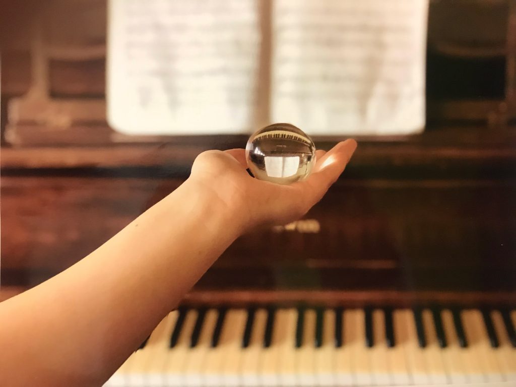 Photographer holding out a clear glass ball on the palm of her hand in front of a piano with sheet music. The piano is flipped upside-down when seen through the glass ball.
